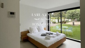 A modern bedroom with a large bed, a thermostat, and a view of a green lawn and chairs outside.