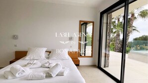 Une chambre d’hôtel avec un grand lit, du linge de lit blanc, une tête de lit en bois, une table de chevet, un miroir et une vue sur des palmiers et une piscine, le tout visible à travers de grandes fenêtres.
