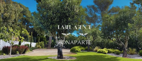 A well-maintained garden with a variety of trees and shrubs, a clear sky, and a paved area in the center.