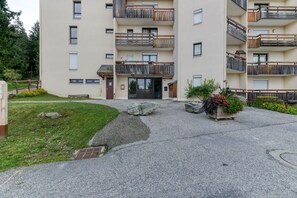 A multi-story building with balconies, a paved entrance area, and a small garden with flowers and rocks. - Apartment 'Les Sapins 1' with Mountain View and Balcony (Les Adrets)