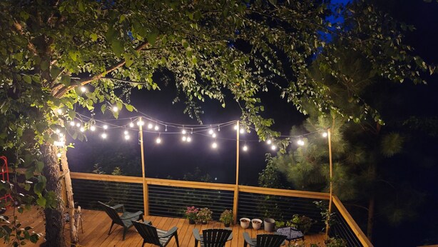 Una terraza de madera con luces colgantes, sillas y plantas en macetas.