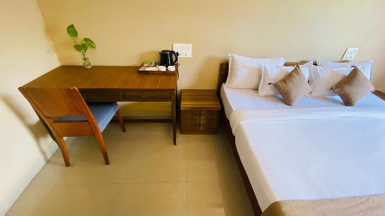Habitación de hotel con escritorio de madera, una silla, cama con ropa de cama blanca y almohadas beige, una mesita de noche pequeña y una planta en maceta sobre el escritorio.
