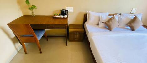 Habitación de hotel con escritorio de madera, una silla, cama con ropa de cama blanca y almohadas beige, una mesita de noche pequeña y una planta en maceta sobre el escritorio.