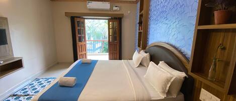 A hotel room with a bed, a ceiling fan, an air conditioning unit, a wooden nightstand, a potted plant, and a view of the outdoors through an open door.