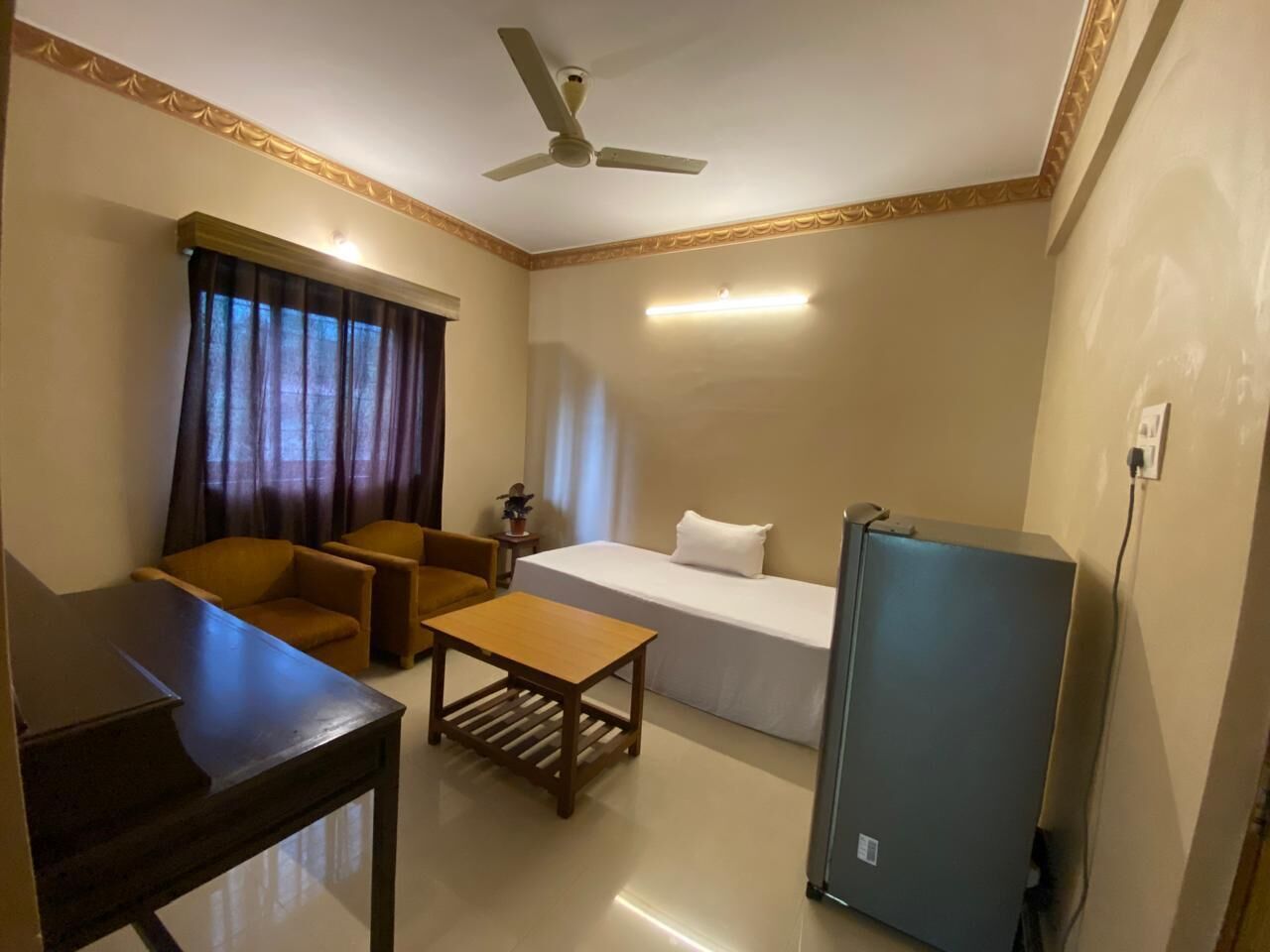 A hotel room with a bed, a desk, a chair, a table, a refrigerator, and a ceiling fan.