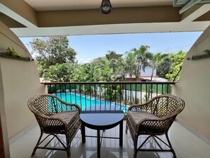 A balcony with two wicker chairs and a small round table, overlooking a swimming pool and lush greenery. - Ashvem Beach Inn (Ashvem)
