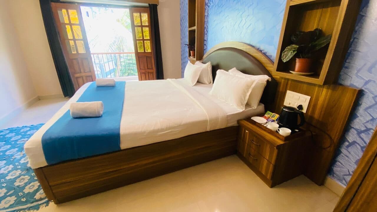 A neatly made bed with white and blue linens, a wooden headboard, and a nightstand with a kettle and cups.