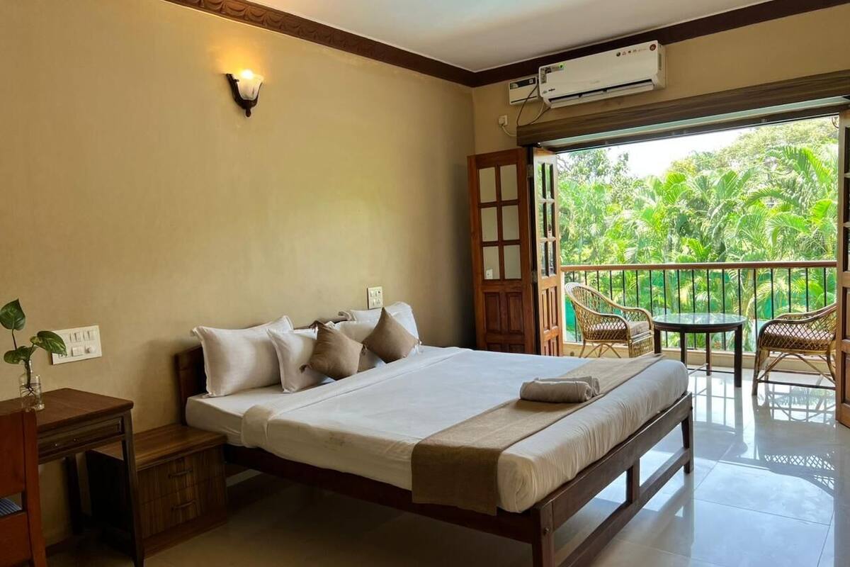 A hotel room with a large bed, a bedside table, a chair, and a balcony with a view of greenery.