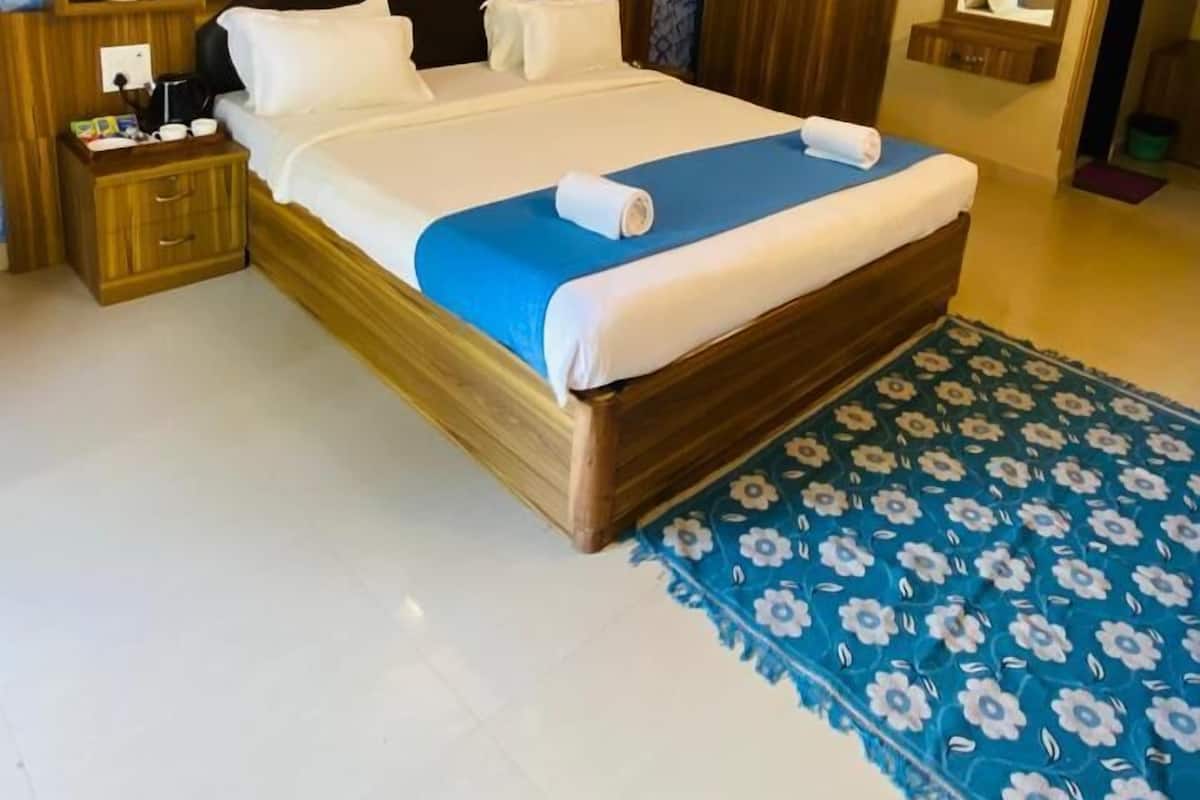 A neatly made bed with a blue bedspread and white pillows, a wooden nightstand with a lamp, a mirror above the bed, and a patterned rug on the floor.