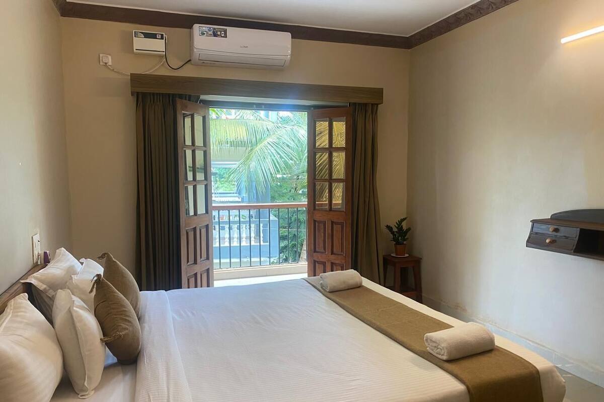 A hotel room with a large bed, a bedside table, an air conditioning unit, a view of the outdoors, and a wall-mounted TV.