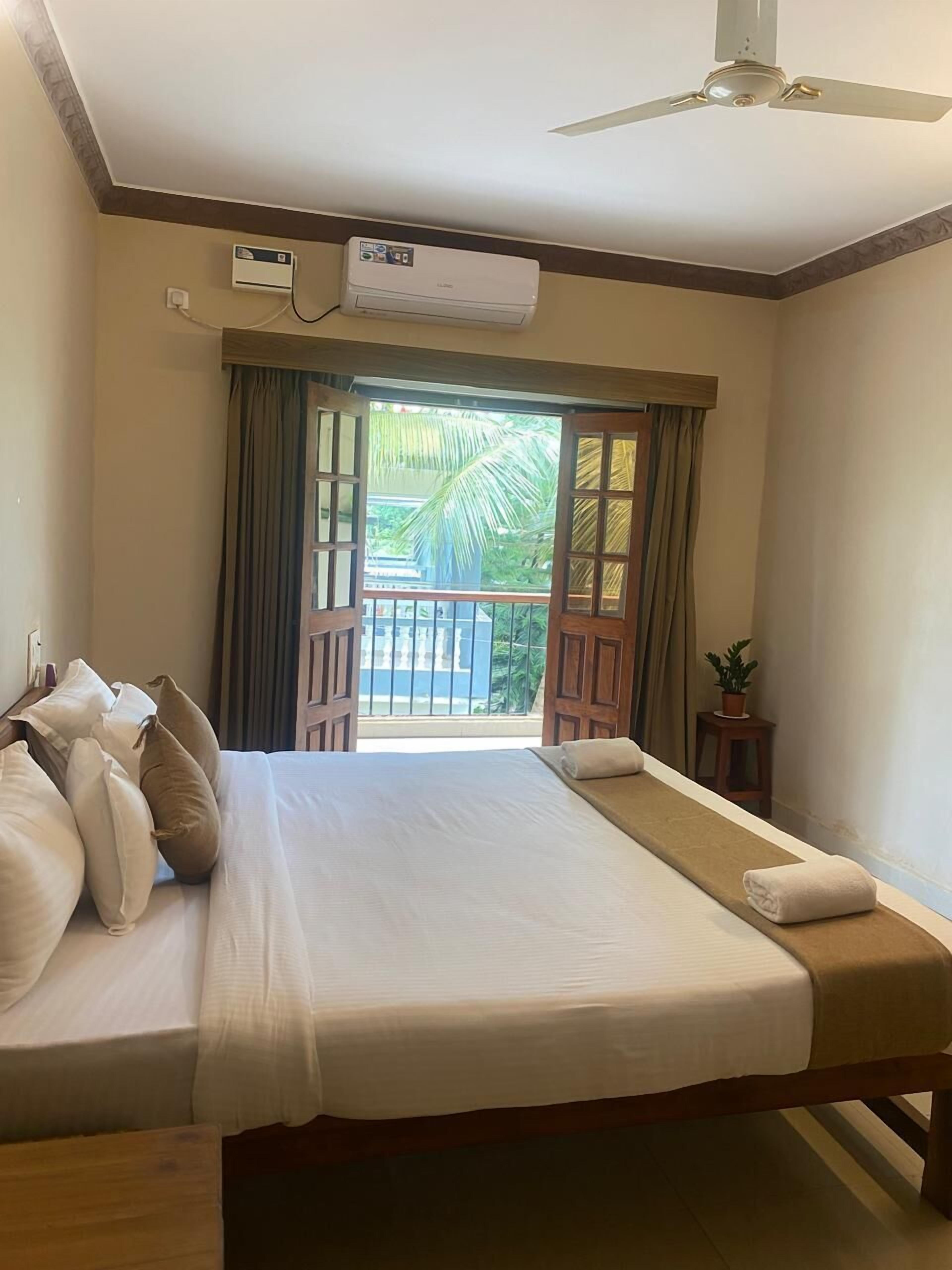 A hotel room with a bed, a ceiling fan, an air conditioning unit, a window with curtains, and a view of a balcony with a palm tree.