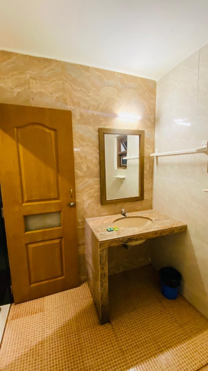 A bathroom with a wooden door, a marble wall, a sink with a mirror, and a blue trash bin.