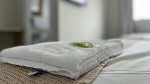 A neatly folded white towel on a textured mat, a bed with white linens, and a wall-mounted TV. - PENHA SOLAR INN (Centro Penha)