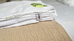 A neatly folded stack of white towels with a green logo on a beige textured ottoman on a bed. - PENHA SOLAR INN (Centro Penha)