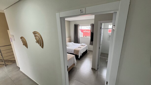 A hotel room with a bed, a bedside table, and a window with curtains. There is a door leading to another room. - PENHA SOLAR INN (Centro Penha)