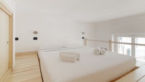 Un dormitorio con piso de madera, una cama con ropa de cama blanca y una ventana.
