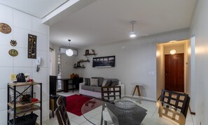 Een moderne woonkamer met een glazen eettafel, een grijze bank en een rood tapijt. Er is een keuken met een fornuis en een koelkast.