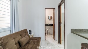 A living room with a sofa, a small side table, and a kitchen area visible through an open door. - Apt Just Minutes From Lagoa da Conceicao - Sis0005 (Florianópolis)
