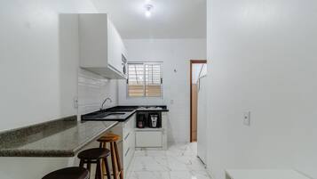Uma cozinha moderna com armários brancos, bancada de granito e fogão embutido. Há três banquetas de madeira e uma máquina de café pequena.