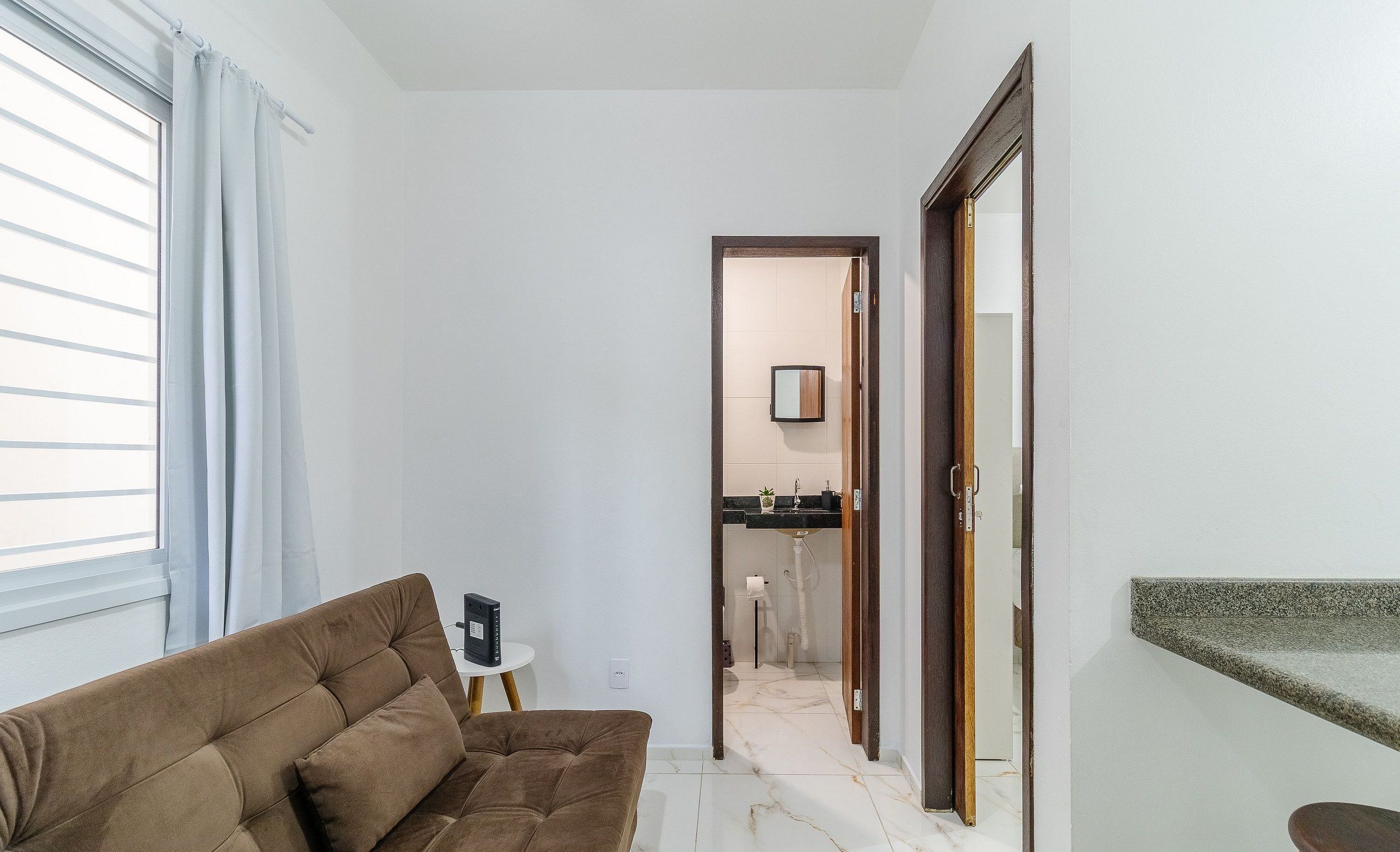 Sala de estar com sofá, uma pequena mesa lateral e cozinha visível através de uma porta aberta.