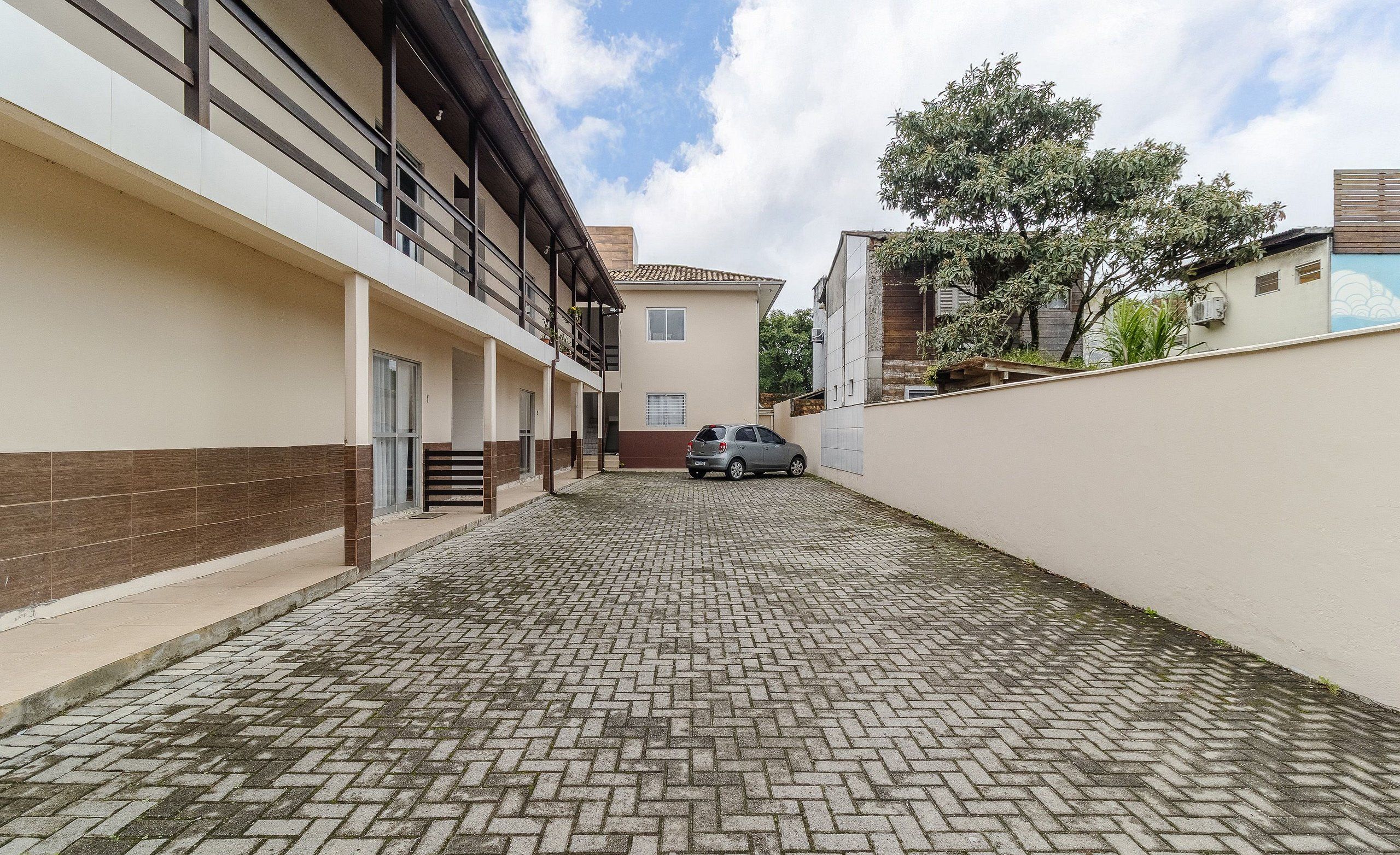 Pátio pavimentado com um carro estacionado ao fundo, rodeado por edifícios com varandas e uma árvore.