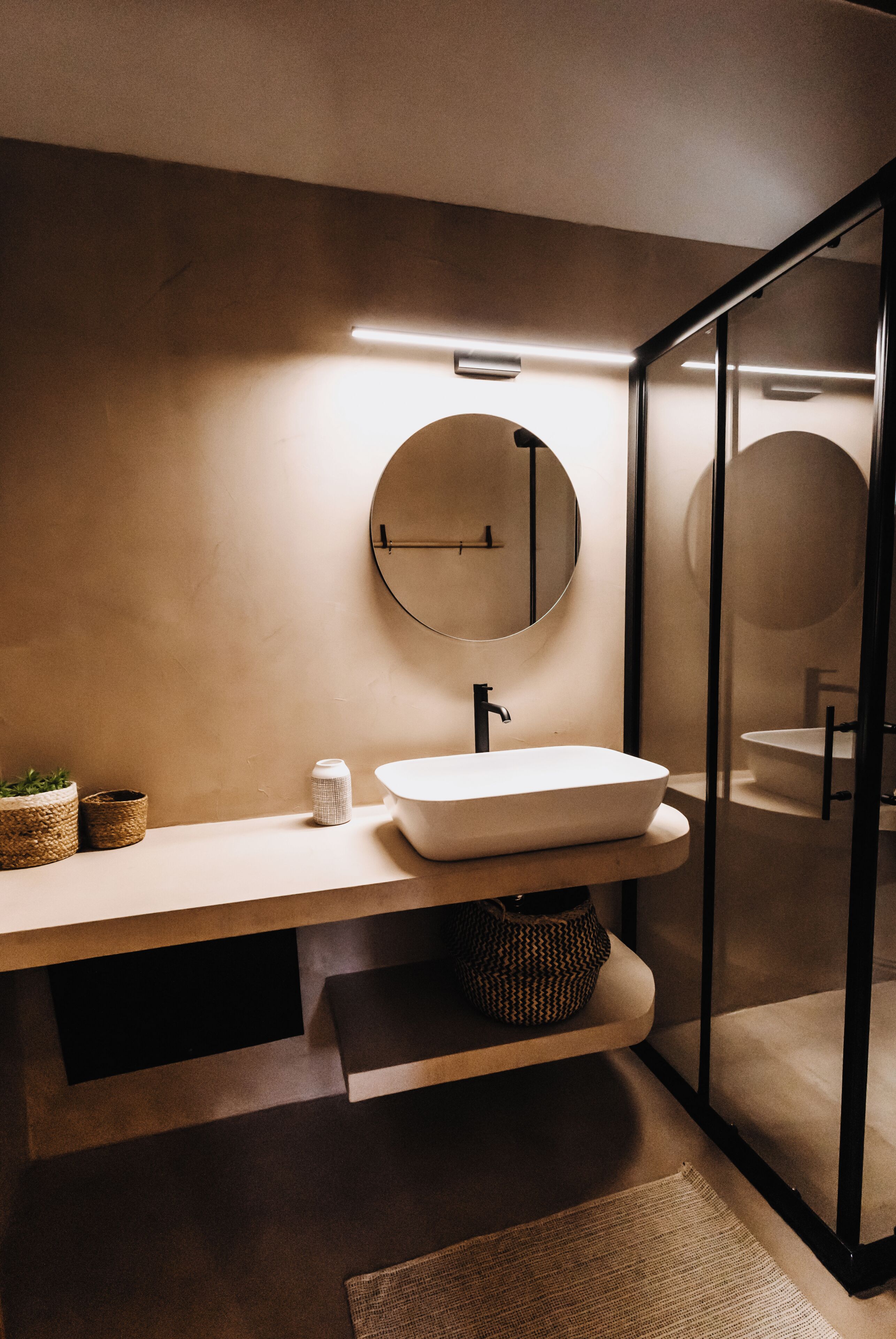 Ein modernes Badezimmer mit rundem Spiegel, Waschbecken in Weiß und einer Duschkabine aus Glas.