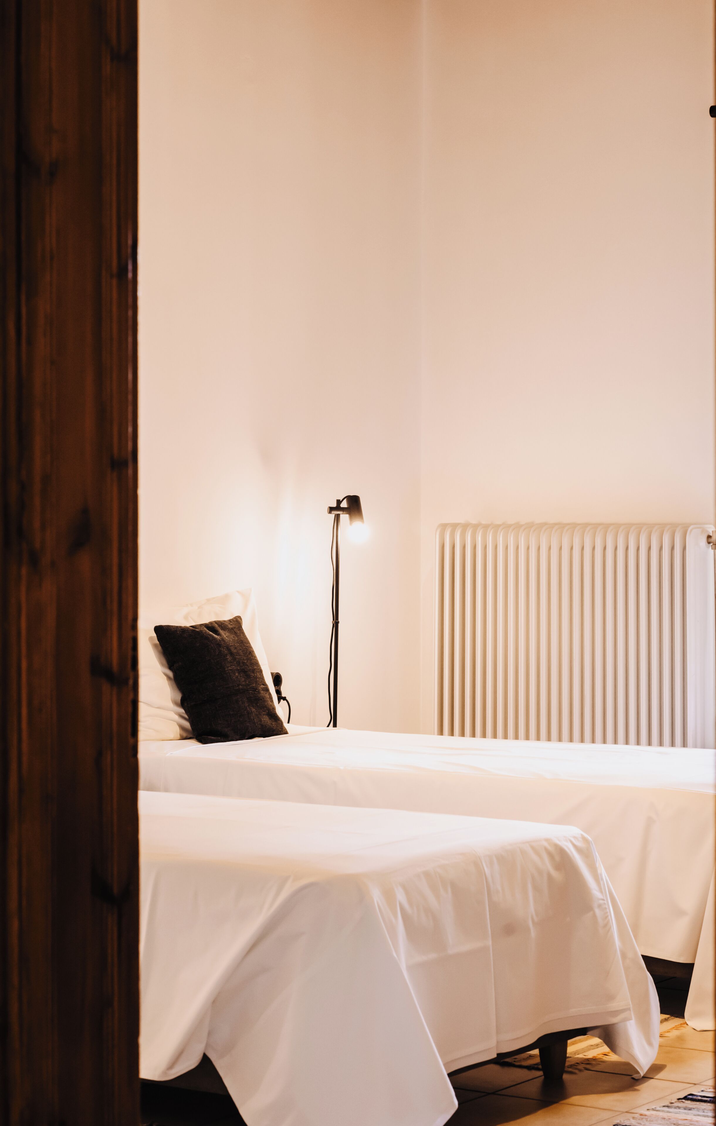 A bedroom with two beds, a bedside lamp, a radiator, and a wooden door.