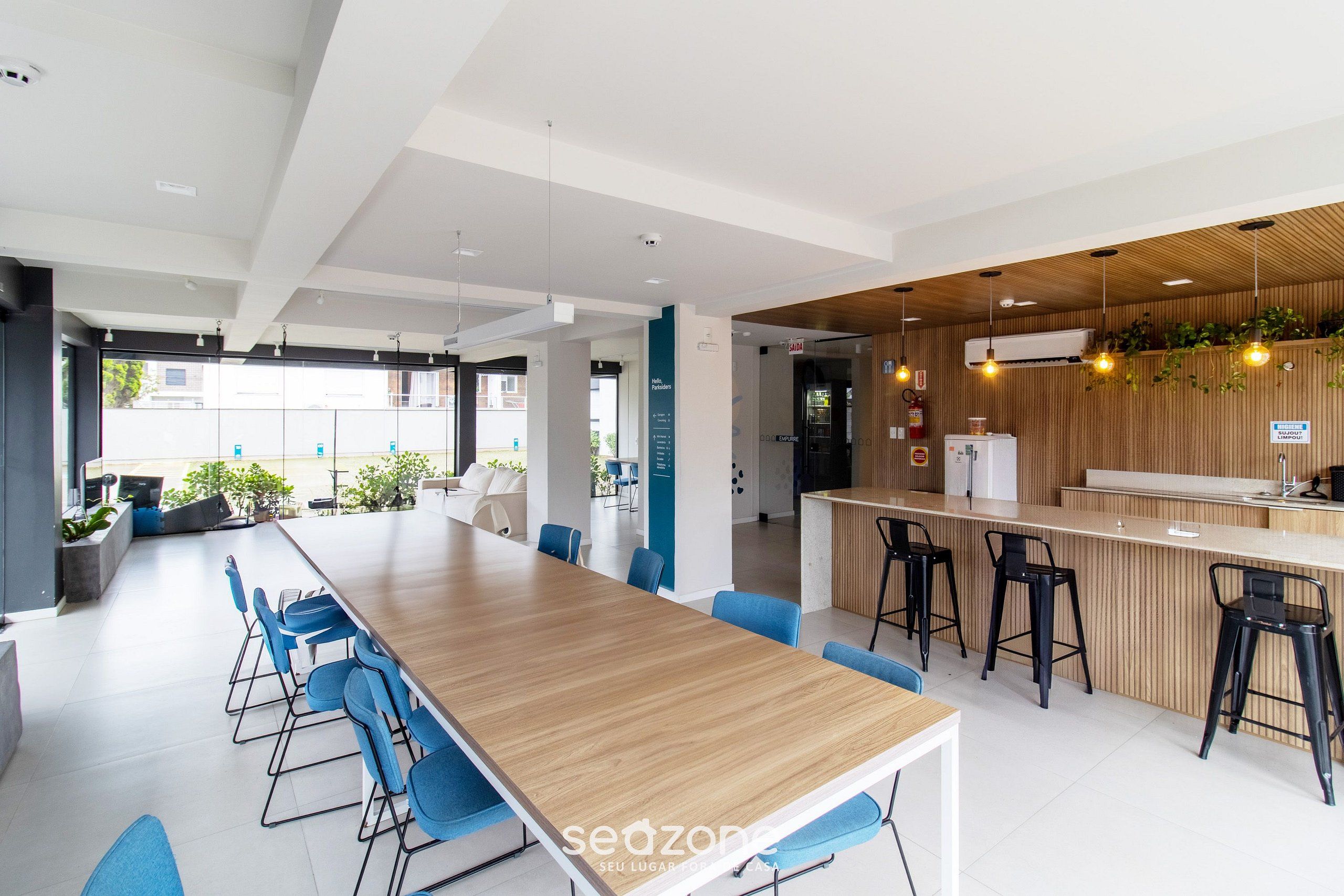 A modern office space with a long wooden table, blue chairs, a bar area with stools, and a view of the outside.
