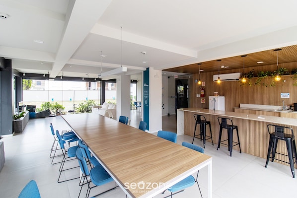 A modern office space with a long wooden table, blue chairs, a bar area with stools, and a view of the outside.