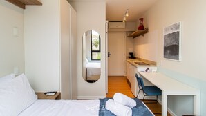 A bedroom with a bed, a desk, a chair, and a small kitchen area. - Modern Studio Just Minutes From the Beach Sid0209 (Florianópolis)