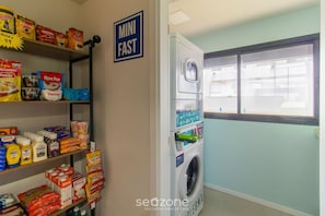 Una sala de lavandería con lavadora y secadora compactas, estantes repletos de productos de limpieza y un cartel que indica el nombre de la sala.