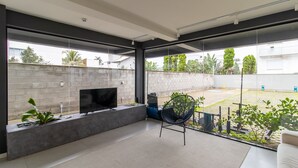 A modern living room with a flat-screen TV on a built-in shelf, a black chair, and a view of the backyard through large windows. - Modern Studio Just Minutes From the Beach Sid0209 (Florianópolis)