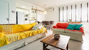 A living room with a yellow sofa, a red and teal sofa, a wooden coffee table, and a dining area with chairs and a table.
