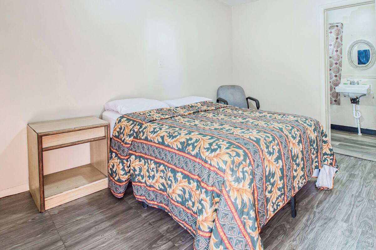 A bedroom with a bed, a bedside table, a chair, and a bathroom with a sink and mirror.