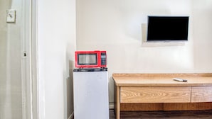 A compact kitchenette with a microwave on top of a mini-fridge, a wooden table with a remote, and a TV mounted on the wall.