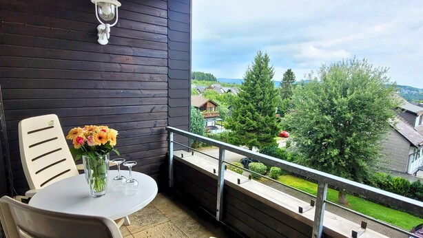 A balcony with a table and chairs, a vase with flowers, and a view of houses and trees. - Apartment With Covered Balcony (Winterberg)
