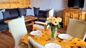 A dining area with a wooden table, chairs, a vase of flowers, and a television on a wooden cabinet. - Apartment With Covered Balcony (Winterberg)