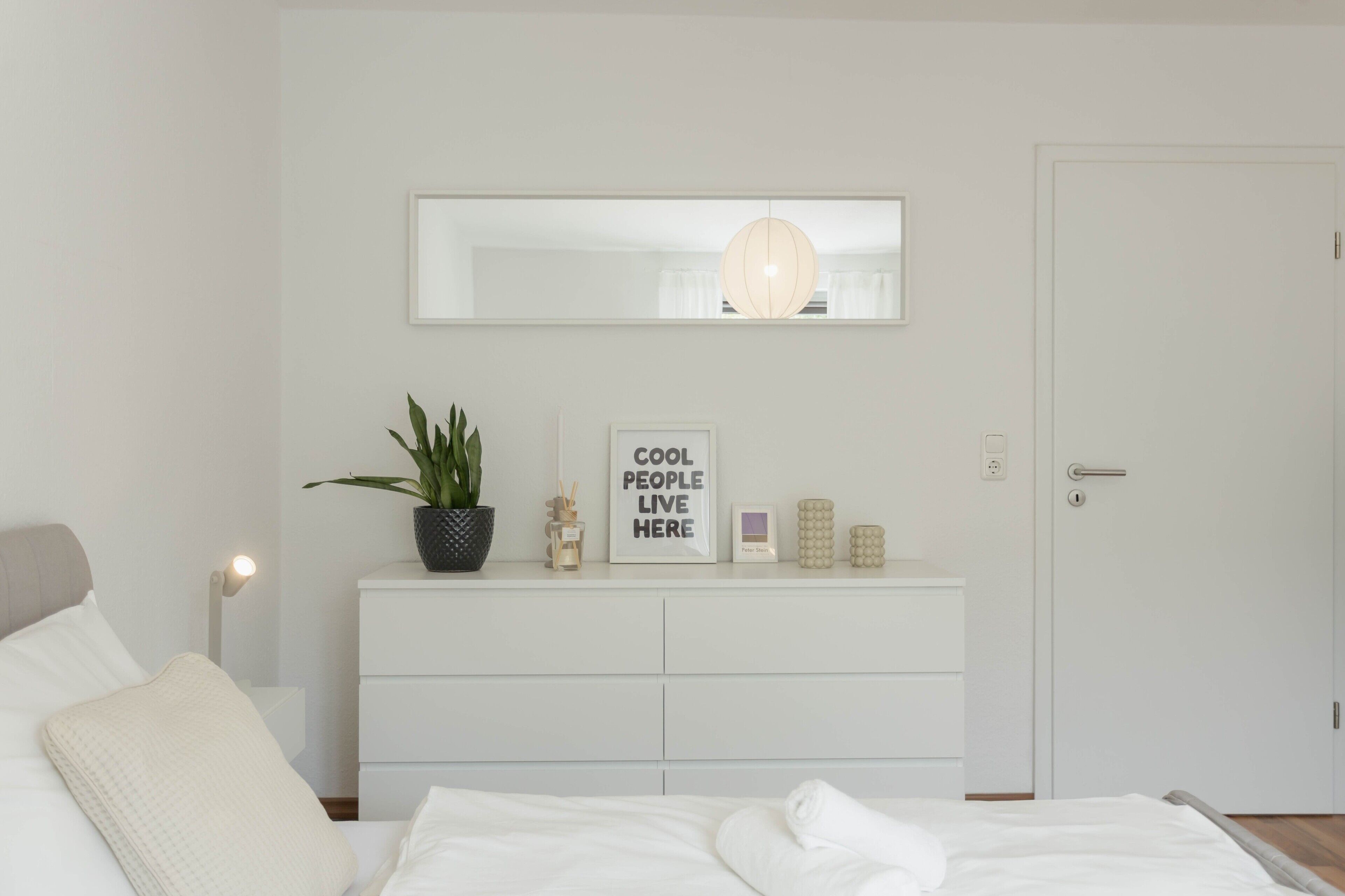 Ein Schlafzimmer mit Bett, Schminktisch, einer Zimmerpflanze, einem Spiegel, einem Schild und einer Tür.