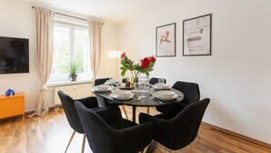 Un comedor con una mesa redonda dispuesta para cuatro personas, rodeada de sillas negras, una planta en maceta con flores rojas y cuadros en la pared.