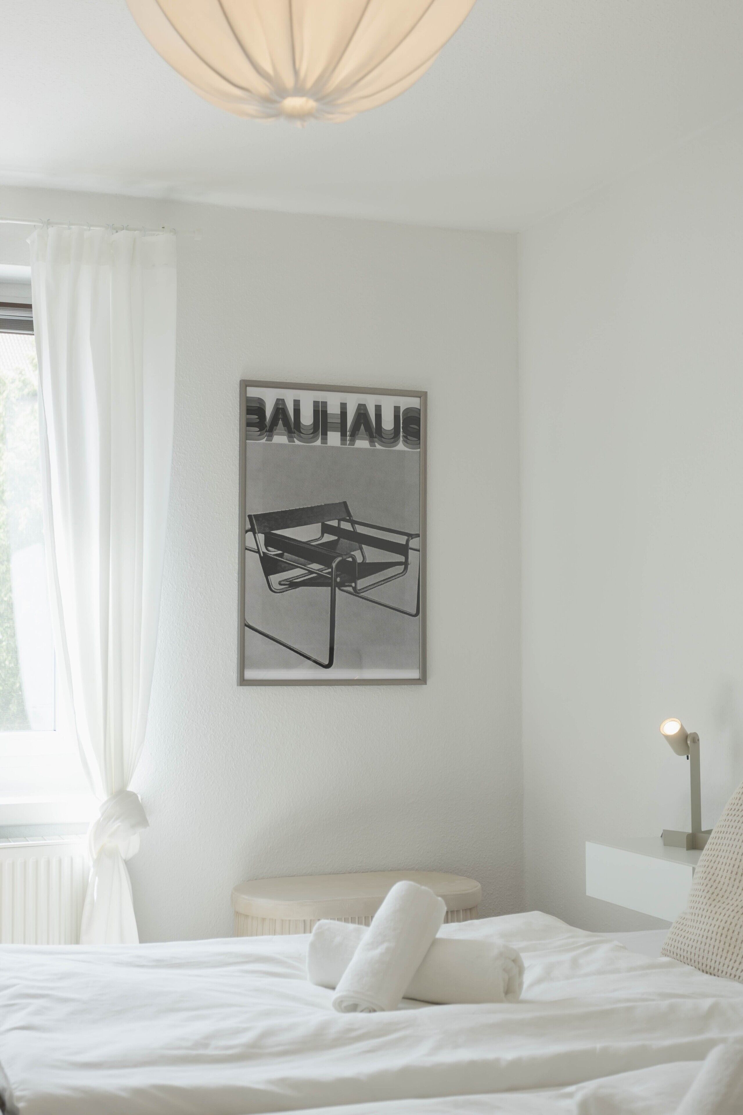 Ein Schlafzimmer mit Bett, Nachttisch, Lampe, einem Fenster mit Vorhängen und einem Bauhaus-Poster an der Wand.