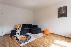 Moderni olohuone, jossa on kulmasohva, sohvapöytä, oranssi jalkarahi ja kuviollinen matto.