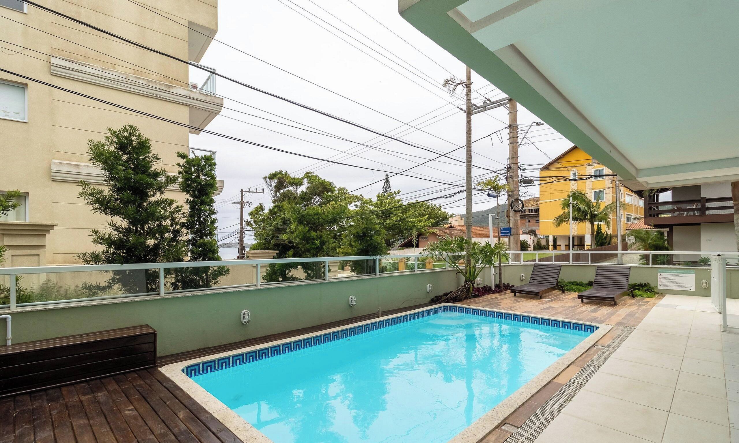 A swimming pool with a wooden deck, surrounded by greenery and a view of neighboring buildings.