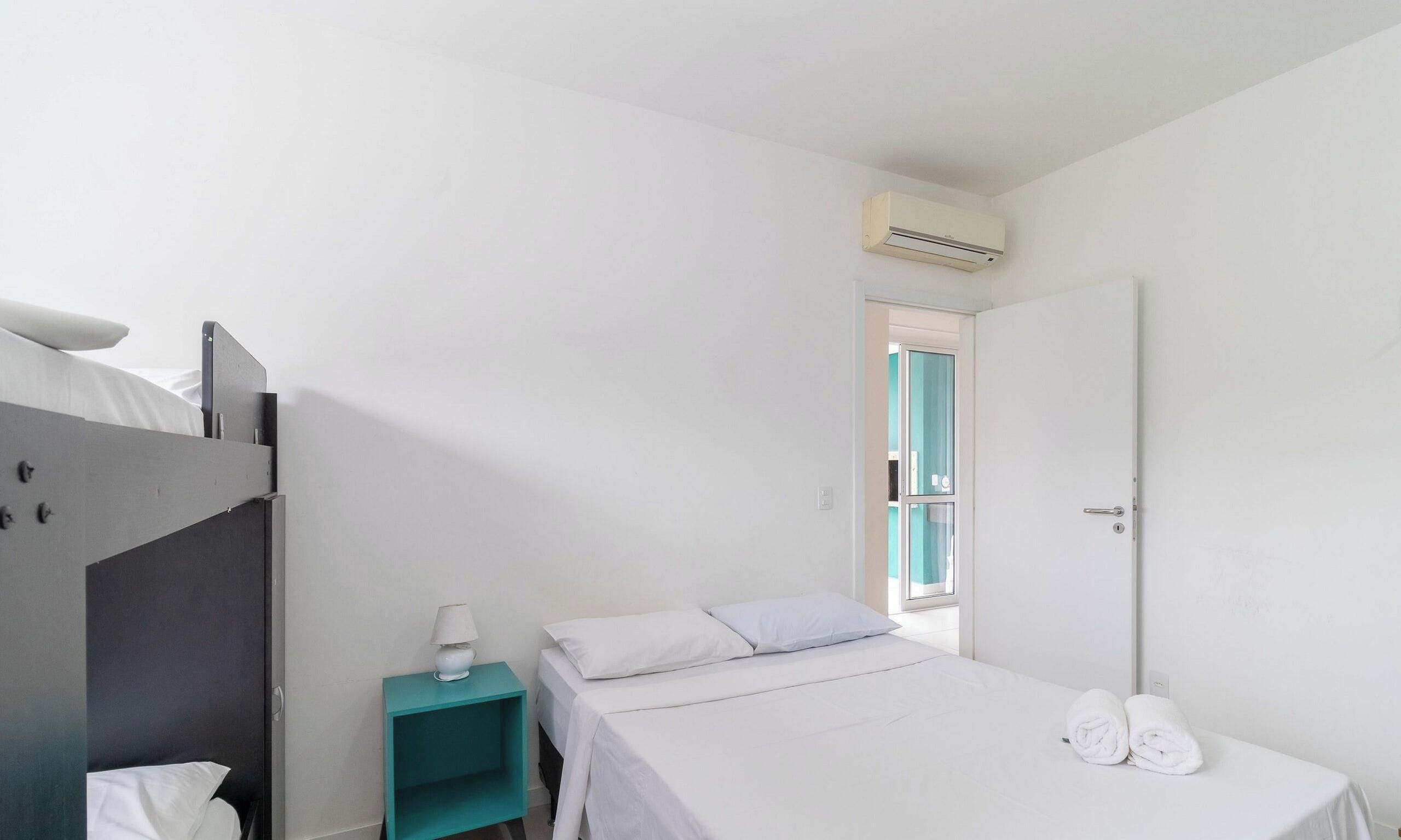 Un dormitorio con una cama litera, una cama individual, una mesita de noche con lámpara y una puerta que da a un balcón.