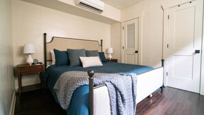 A bedroom with a bed, two bedside tables, a nightstand, a window with blinds, and a door.