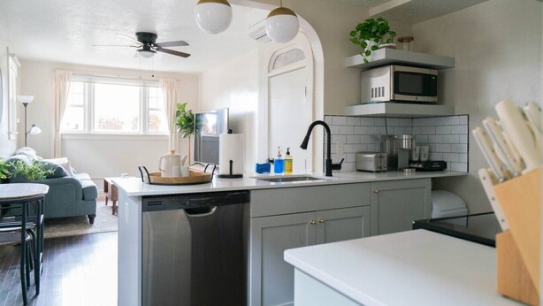 A modern kitchen with grey cabinets, a white countertop, a built-in microwave, and a dishwasher. There is a dining area with a table and chairs, and a ceiling fan above.