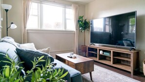 A living room with a sofa, a coffee table, a TV on a wooden stand, a lamp, and a window with curtains.