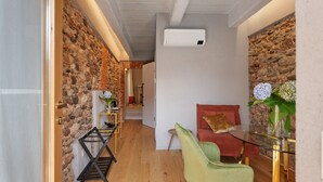 Habitació amb paret de pedra, un sofà vermell, una cadira verda, taula de vidre i aire condicionat penjat.
