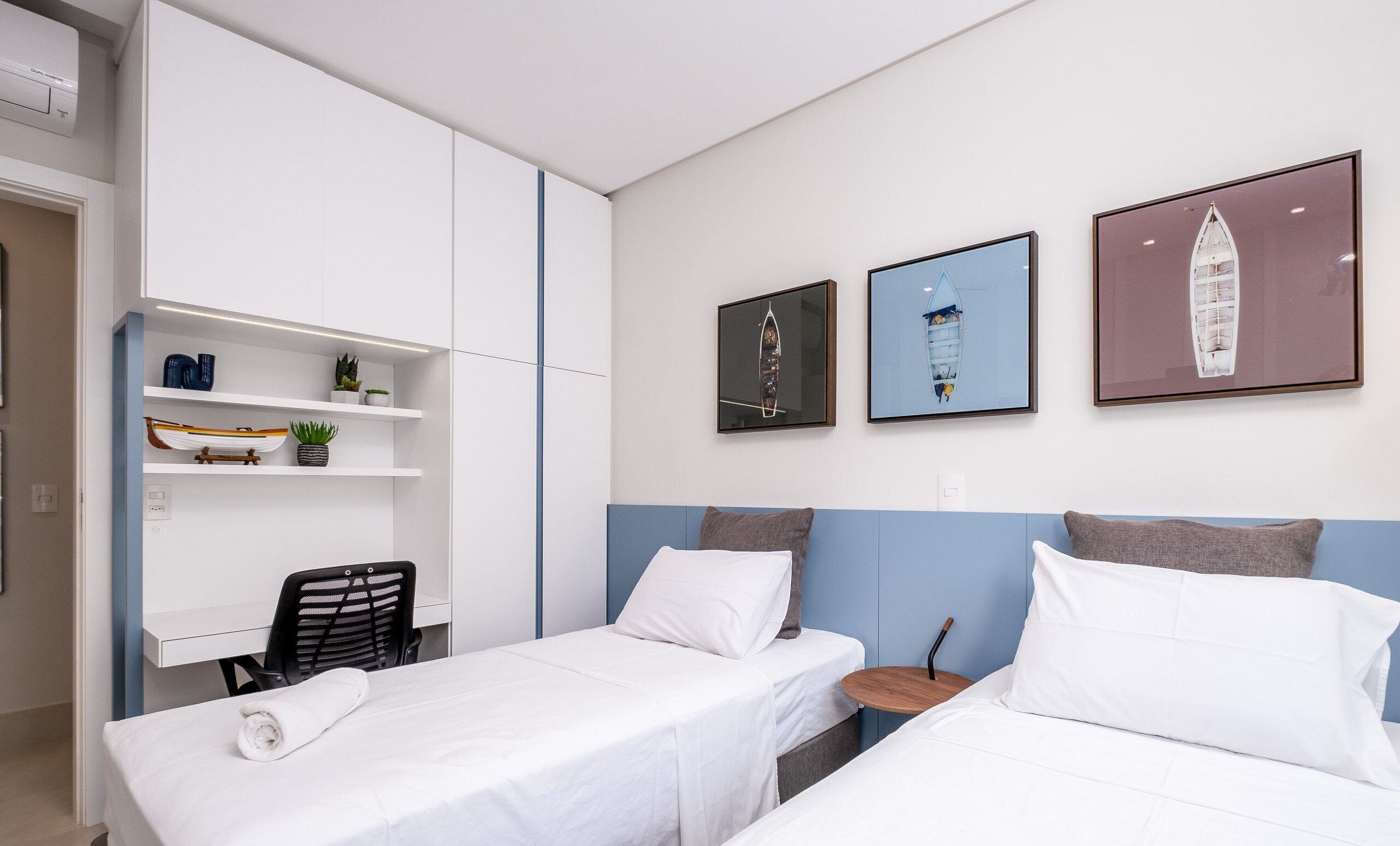 A hotel room with two beds, a desk with a chair, a shelf with decorative items, and three framed pictures on the wall.