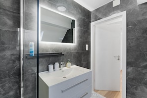 Ein modernes Badezimmer mit einem weißen Waschbecken, einem Spiegel mit integrierter Beleuchtung und dunklen Marmorwänden.