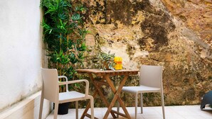 Un pequeño espacio al aire libre con dos sillas blancas y una mesa de madera, frente a una pared de piedra con vegetación.